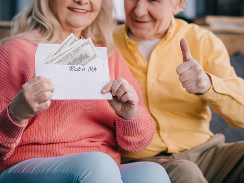 cropped view of senior couple showing thumb up sign while holding envelope with 'roth ira' lettering