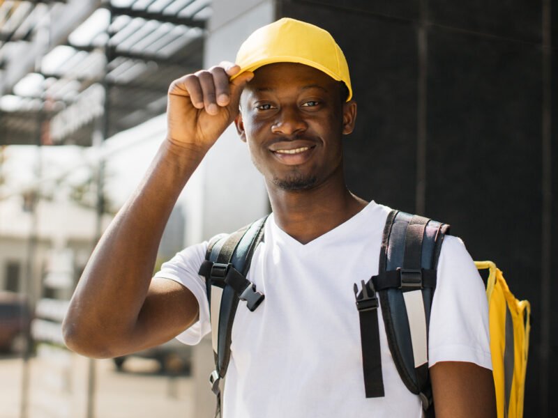 Food delivery, portrait of courier african man with yellow backpack and cap. Quick service, work concept for foreigners, simple profession.