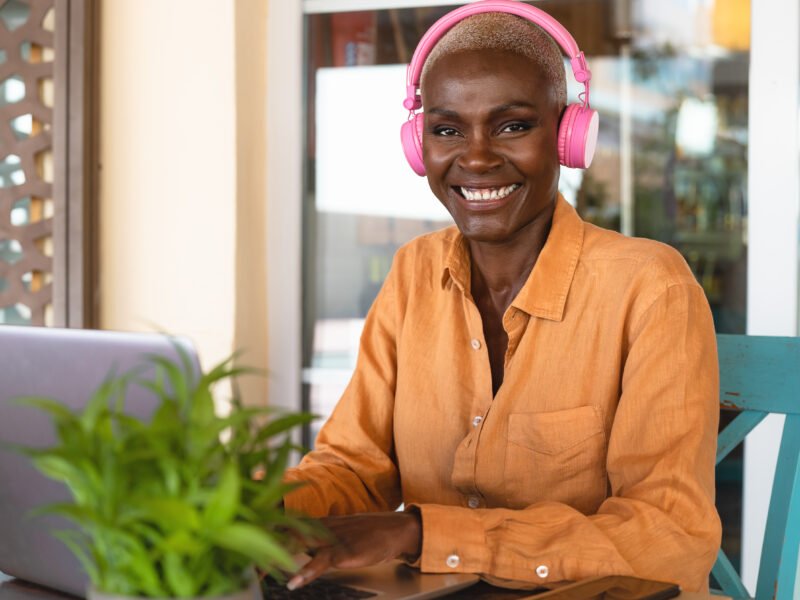 Smiling African woman using laptop and listening music with headphones in bar restaurant - Digital nomad and freelance lifestyle concept